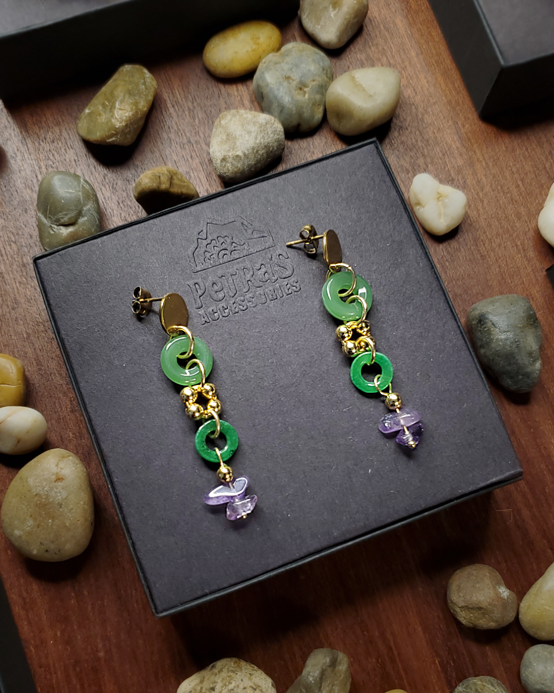 Circle gold stainless steel earring studs with jade circle beads, gold beads and amethyst stone beads. Handcrafted earrings inside a PeTRa's ACCESSORIES embossed logo black jewelry box. Brown wood table and loose rocks background.