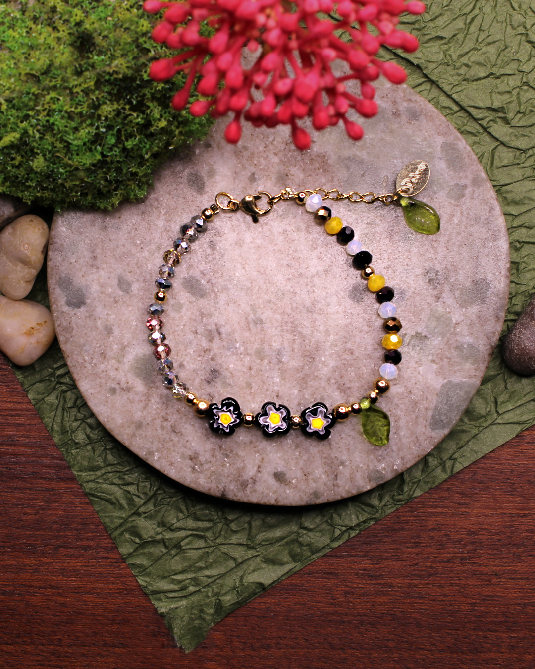 Colorful bracelet made of faceted crystal beads in colors: yellow, white, gray, black and black  millefiori beads. Bracelet on top of a gray stone display. Natural red flower, rocks, green and brown background.