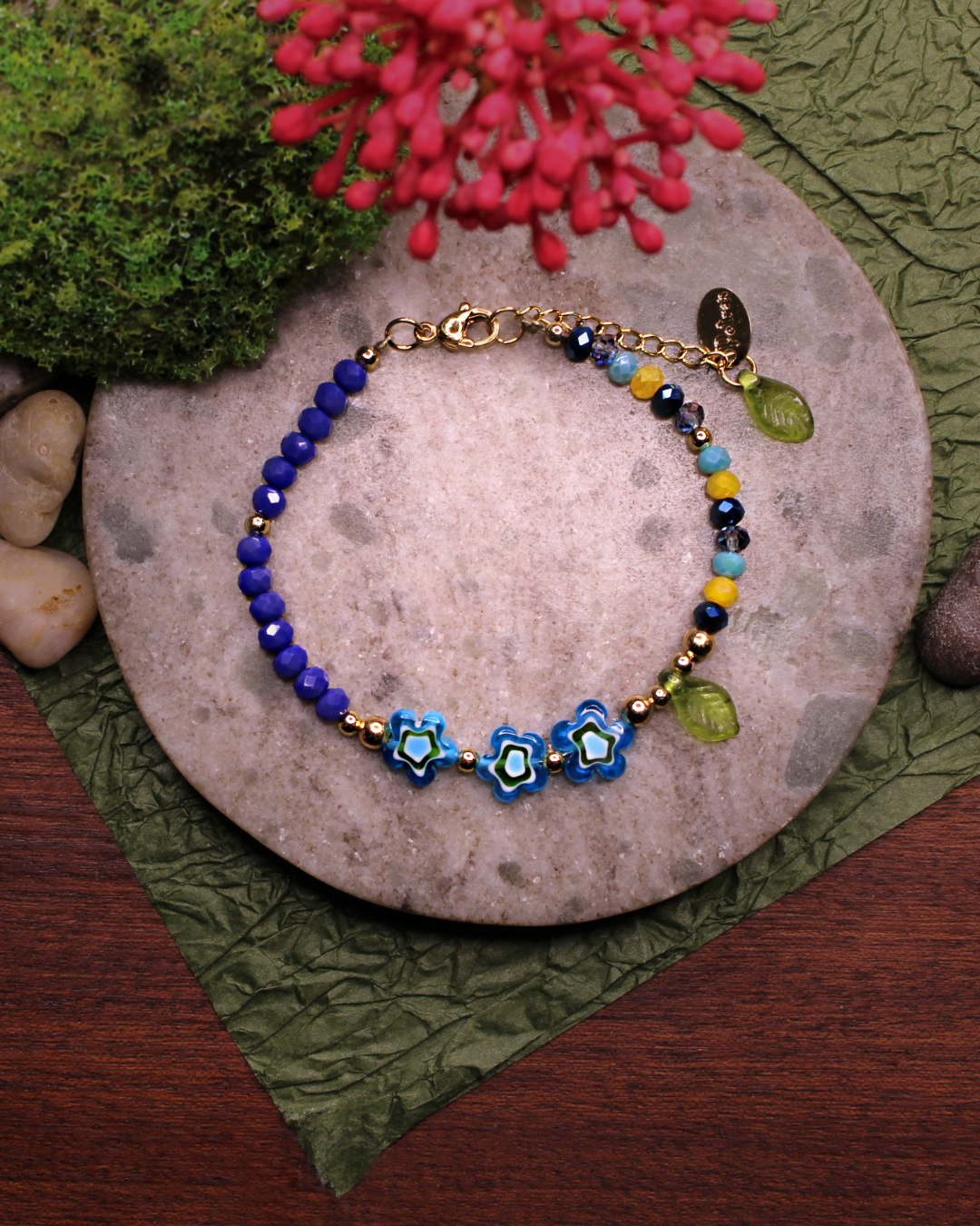 Colorful bracelet made of faceted crystal beads in colors: yellow, royal blue, dark blue, transparent blue, light blue and millefiori beads. Bracelet on top of a gray stone display. Natural red flower, rocks, green and brown background.