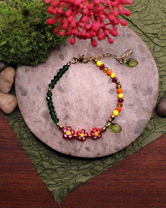 Colorful bracelet made of faceted crystal beads in colors: yellow, red, amber, dark green and millefiori beads. Bracelet on top of a gray stone display. Natural red flower, rocks, green and brown background.