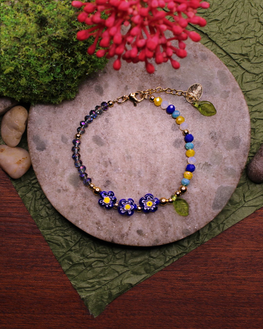 Colorful bracelet made of faceted crystal beads in colors: yellow, white, royal blue, sky blue, transparent and millefiori beads. Bracelet on top of a gray stone display. Natural red flower, rocks, green and brown background.
