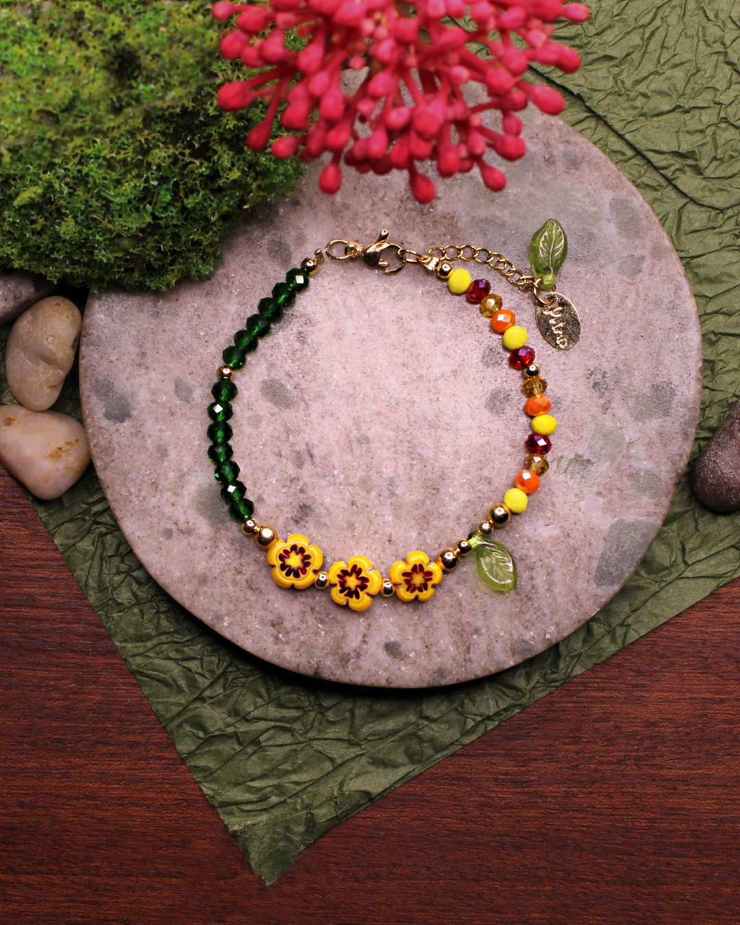Colorful bracelet made of faceted crystal beads in colors: yellow, red, amber, dark green and millefiori beads. Bracelet on top of a gray stone display. Natural red flower, rocks, green and brown background.