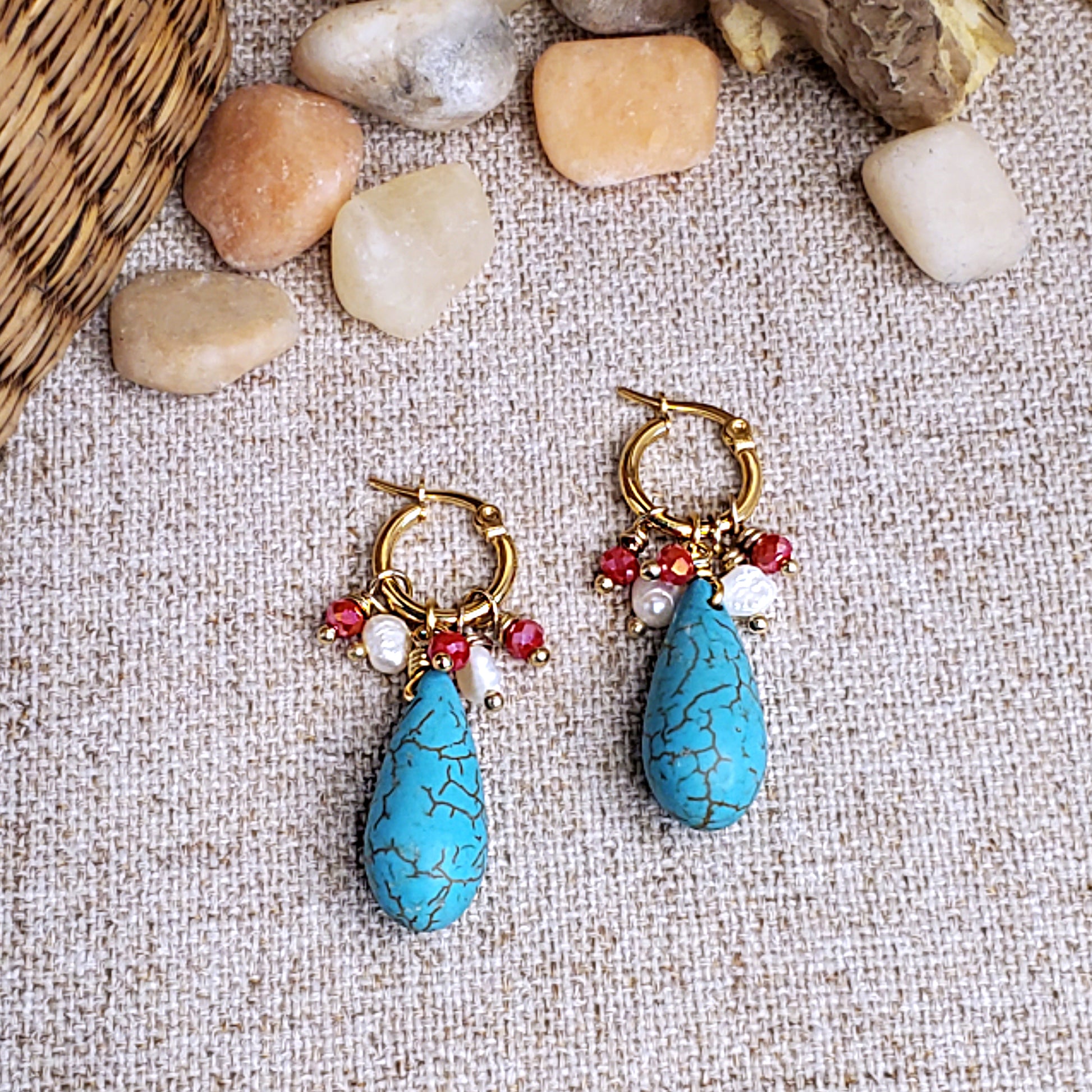 Gold stainless steel earring hoops with red crystals, freshwater pearls and howlite teardrop dangling bead, on a linen cloth background. 