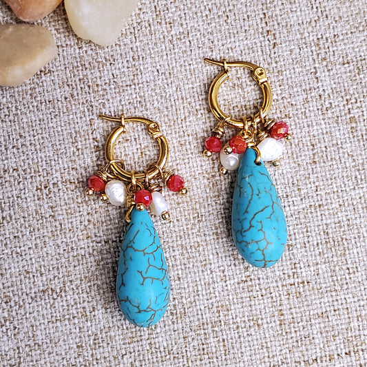 Gold stainless steel earring hoops with red crystals, freshwater pearls and howlite teardrop dangling bead, on a linen cloth background. 