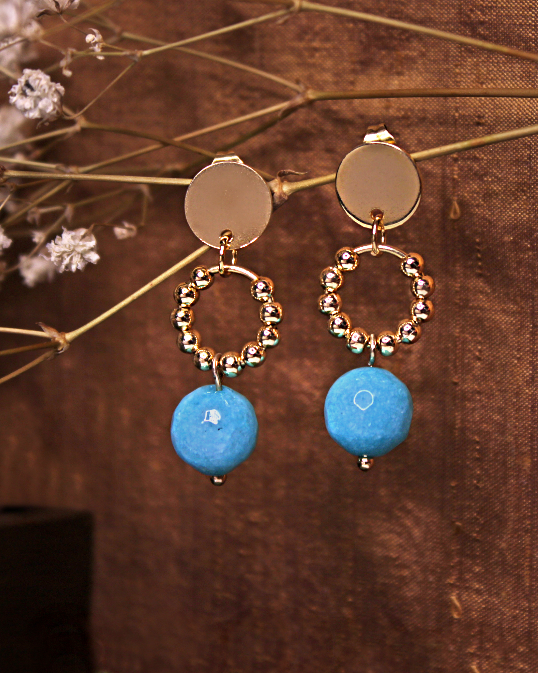 Circle stainless steel dangling stud earrings with gold beads and faceted blue bead, in front of an vintage gold silk fabric and baby breath flowers.