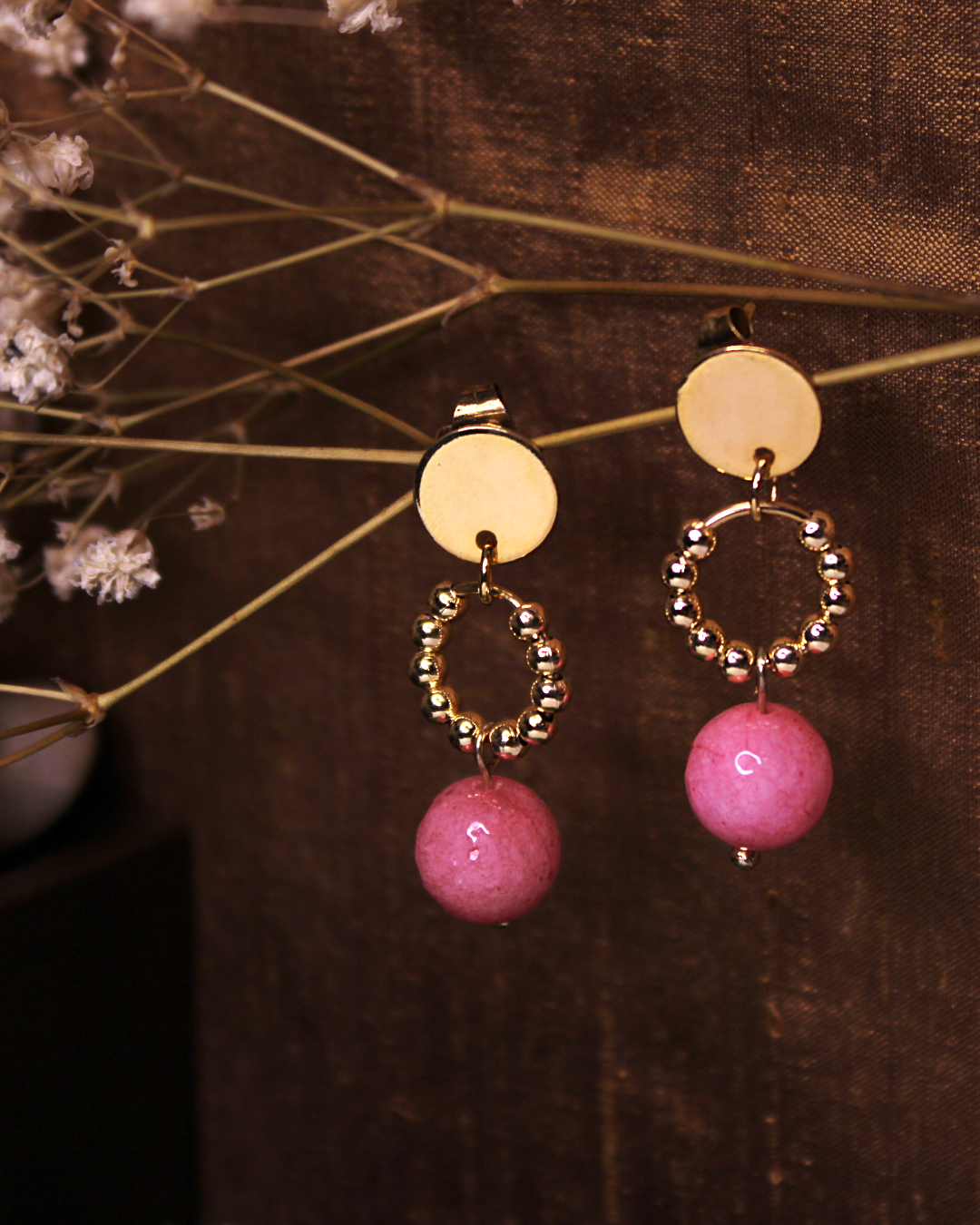 Circle stainless steel dangling stud earrings with gold beads and faceted pink bead, in front of an vintage gold silk fabric and baby breath flowers.